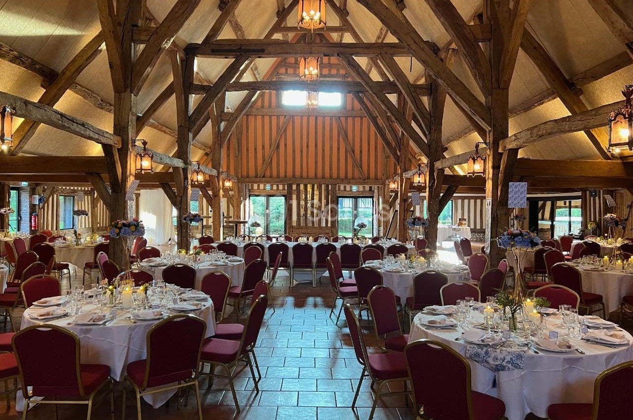 Salle de réception rustique avec poutres apparentes, tables rondes dressées et chaises rouges disposées pour un événement.