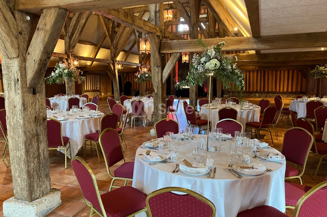Salle de réception avec tables rondes dressées, nappes blanches, chaises rouges et poutres apparentes en bois.