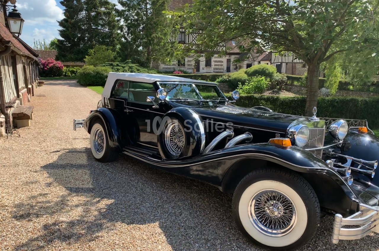 Voiture ancienne noire avec toit blanc garée sur une allée en gravier, entourée de verdure et de bâtiments à colombages.