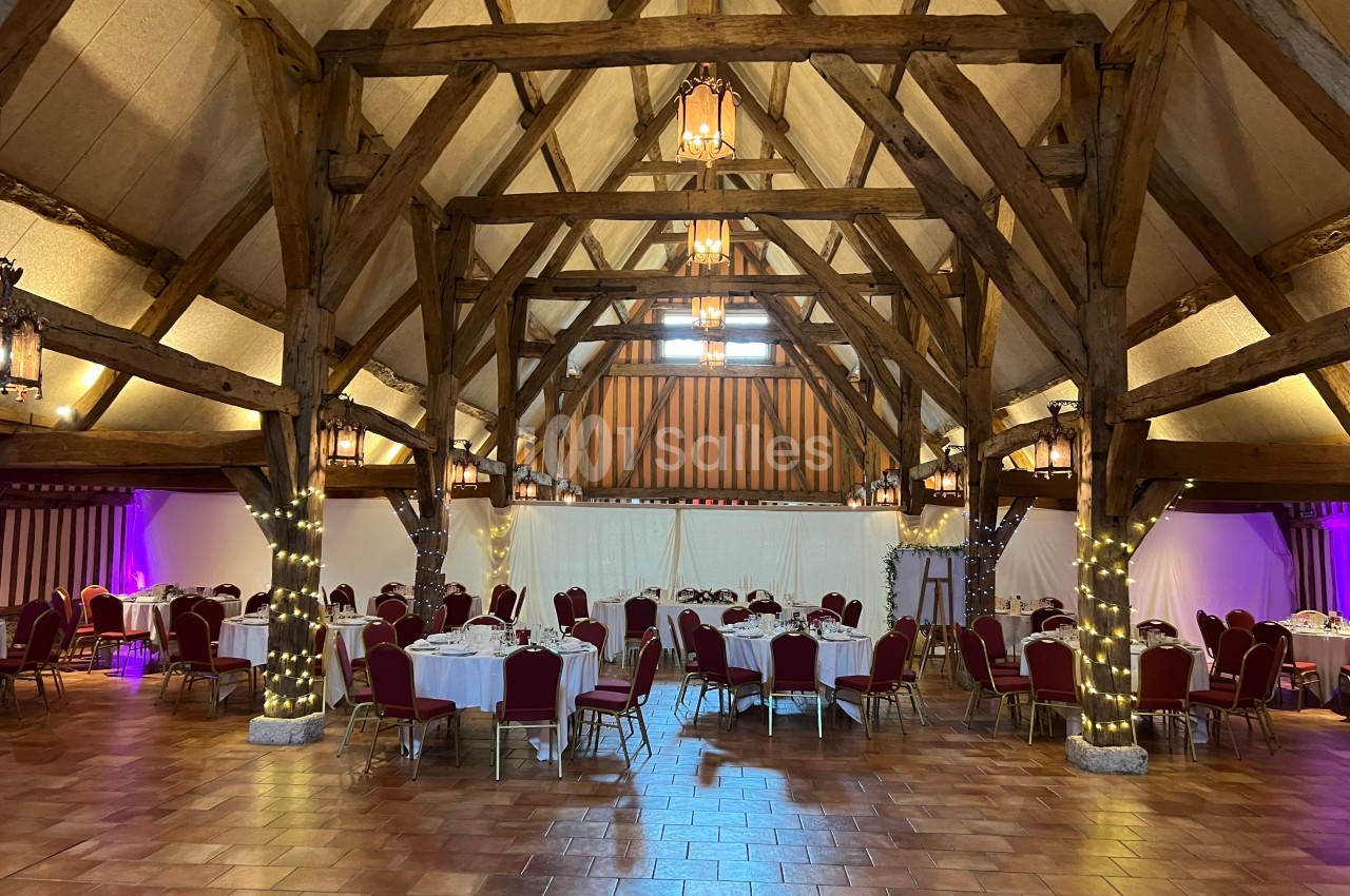Salle de réception avec charpente en bois apparente, tables rondes dressées et chaises rouges sur un sol en carrelage.