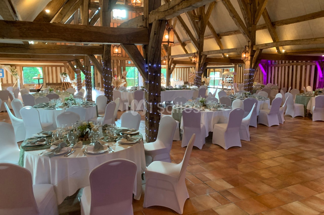 Salle de réception rustique avec poutres apparentes, tables rondes dressées et chaises blanches, éclairage chaleureux.