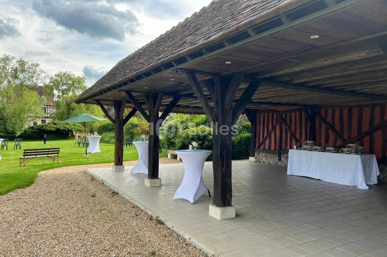 Terrasse couverte en bois avec tables hautes drapées de blanc, donnant sur un jardin verdoyant par temps nuageux.