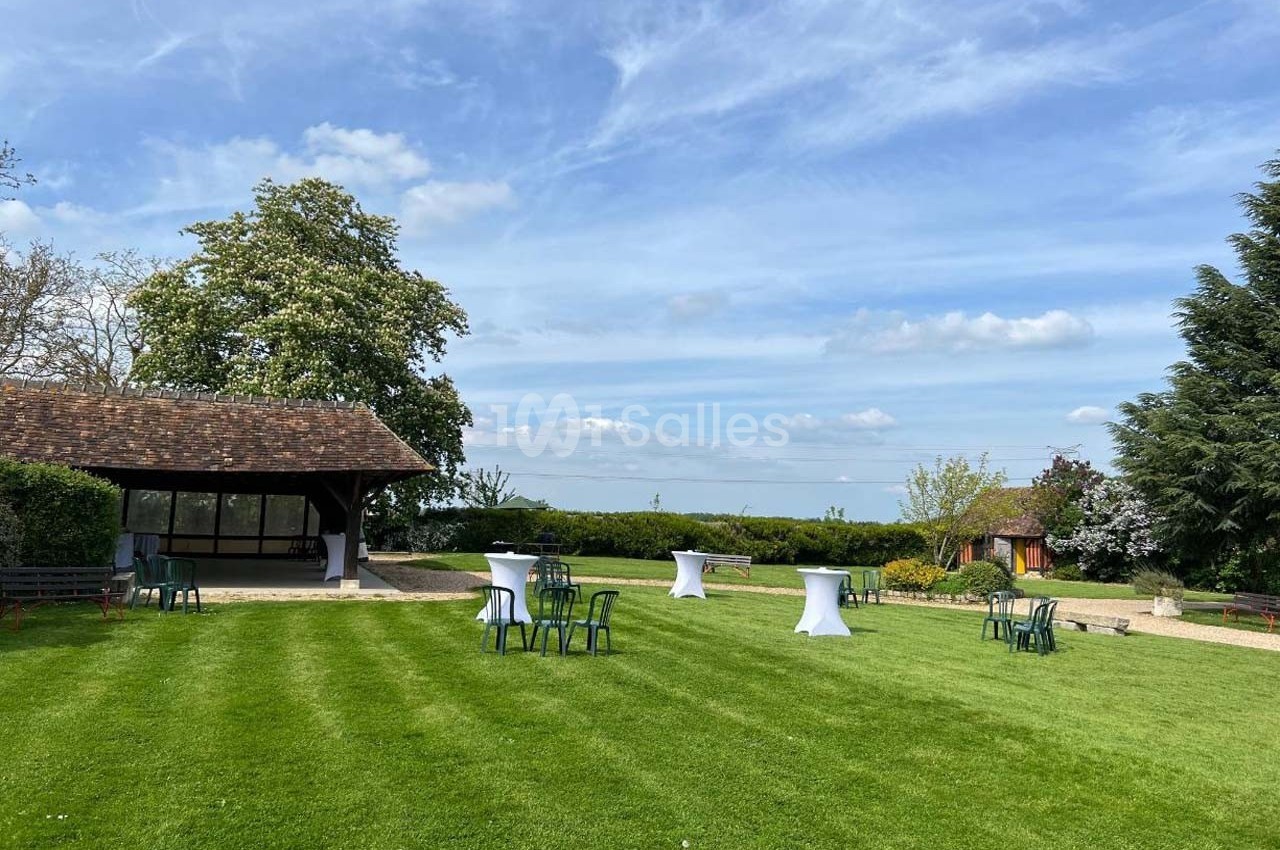 Pelouse verdoyante avec tables hautes et chaises, entourée d'arbres et d'une petite structure en bois sous un ciel dégagé.
