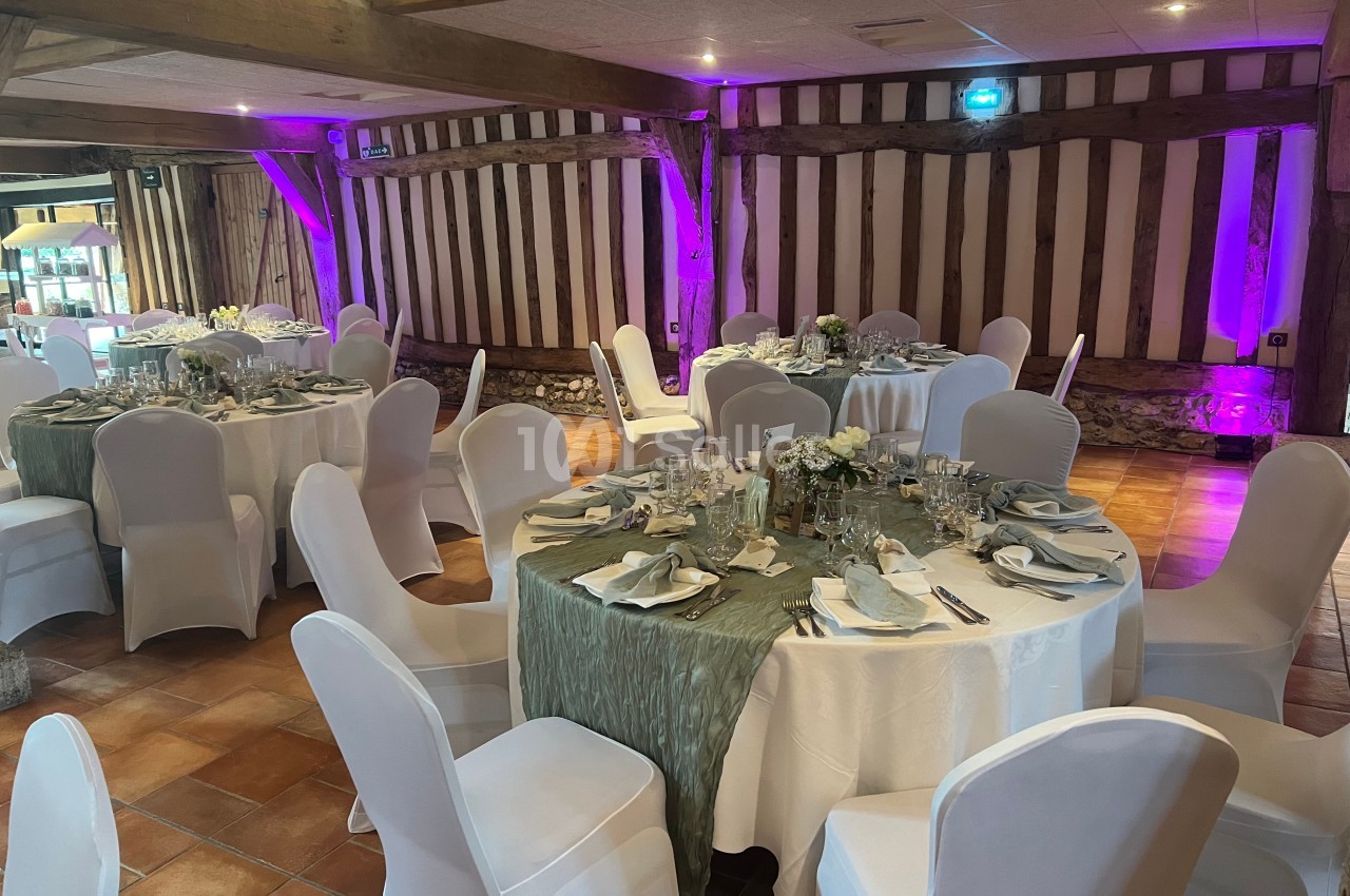 Salle de réception avec tables rondes dressées, nappes blanches et décorations florales, éclairage violet en arrière-plan.