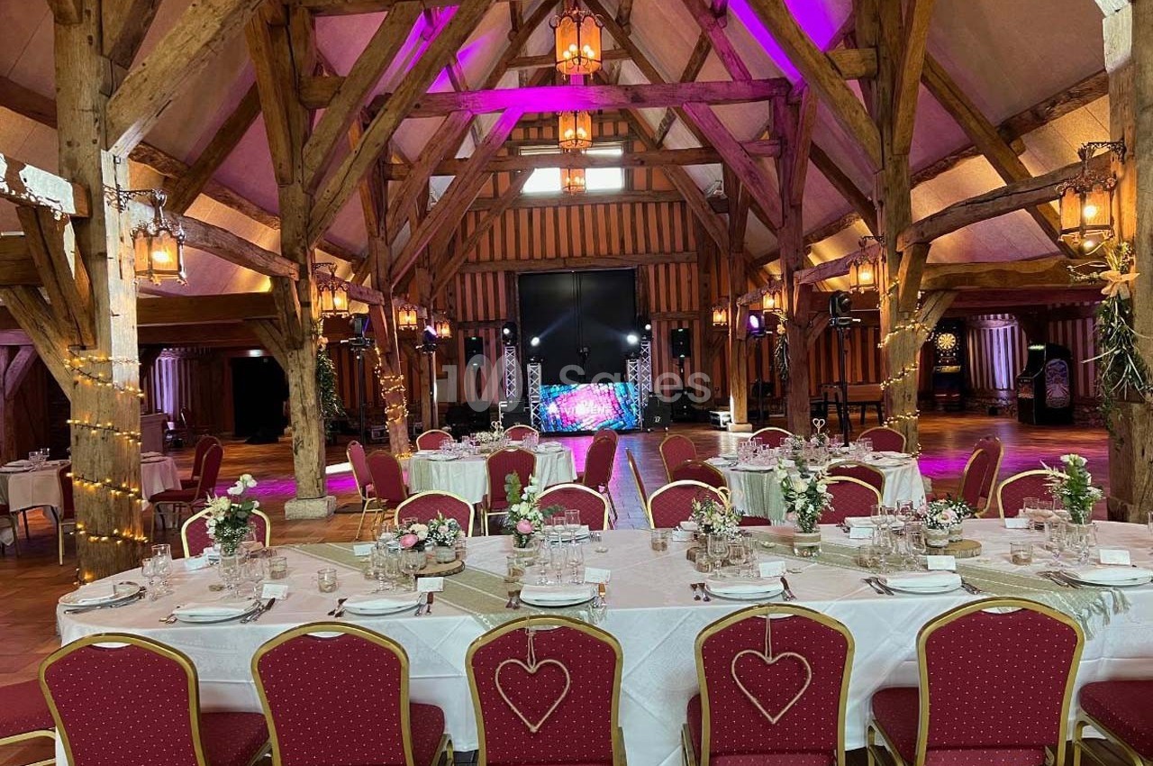 Salle de réception avec poutres apparentes, tables décorées pour un événement et éclairage coloré en arrière-plan.