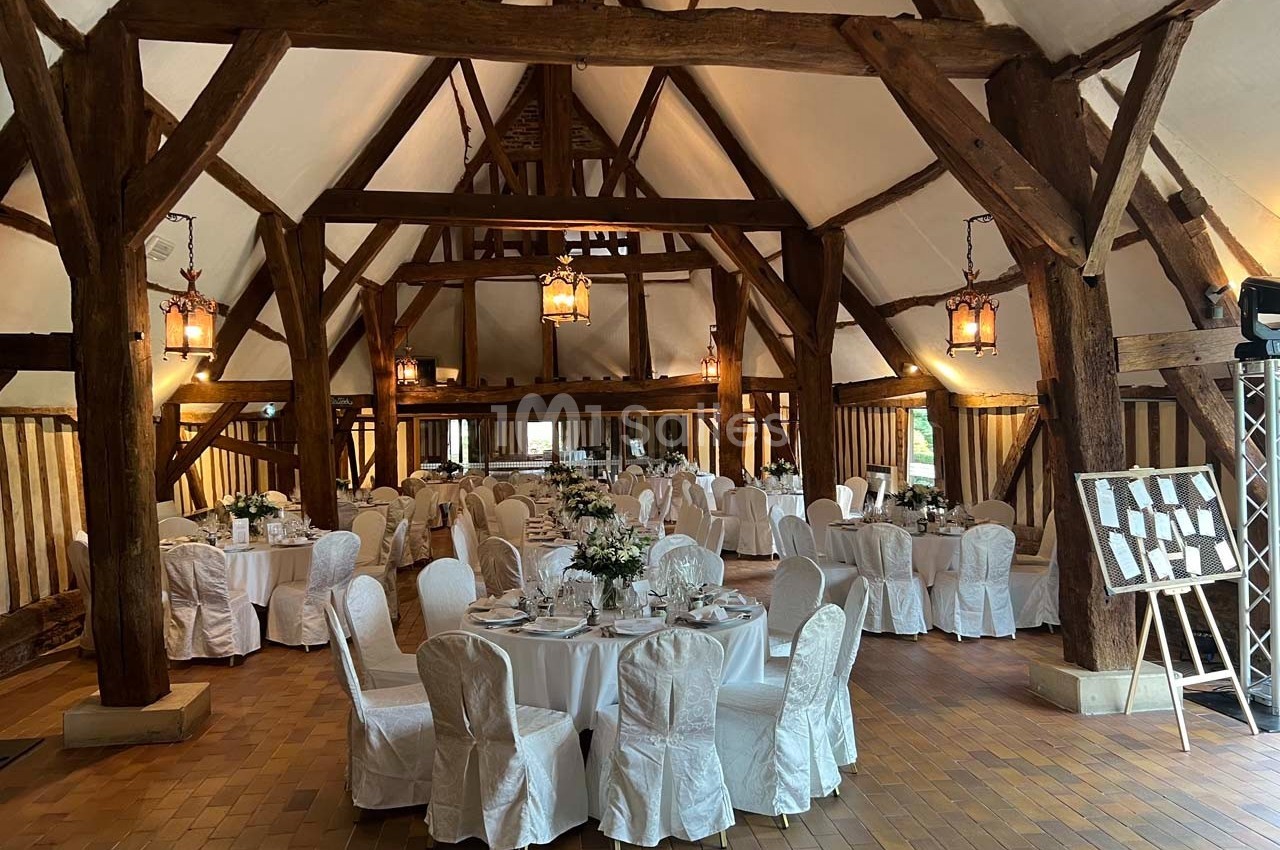 Salle de réception rustique avec poutres en bois apparentes, tables rondes dressées et chaises blanches.