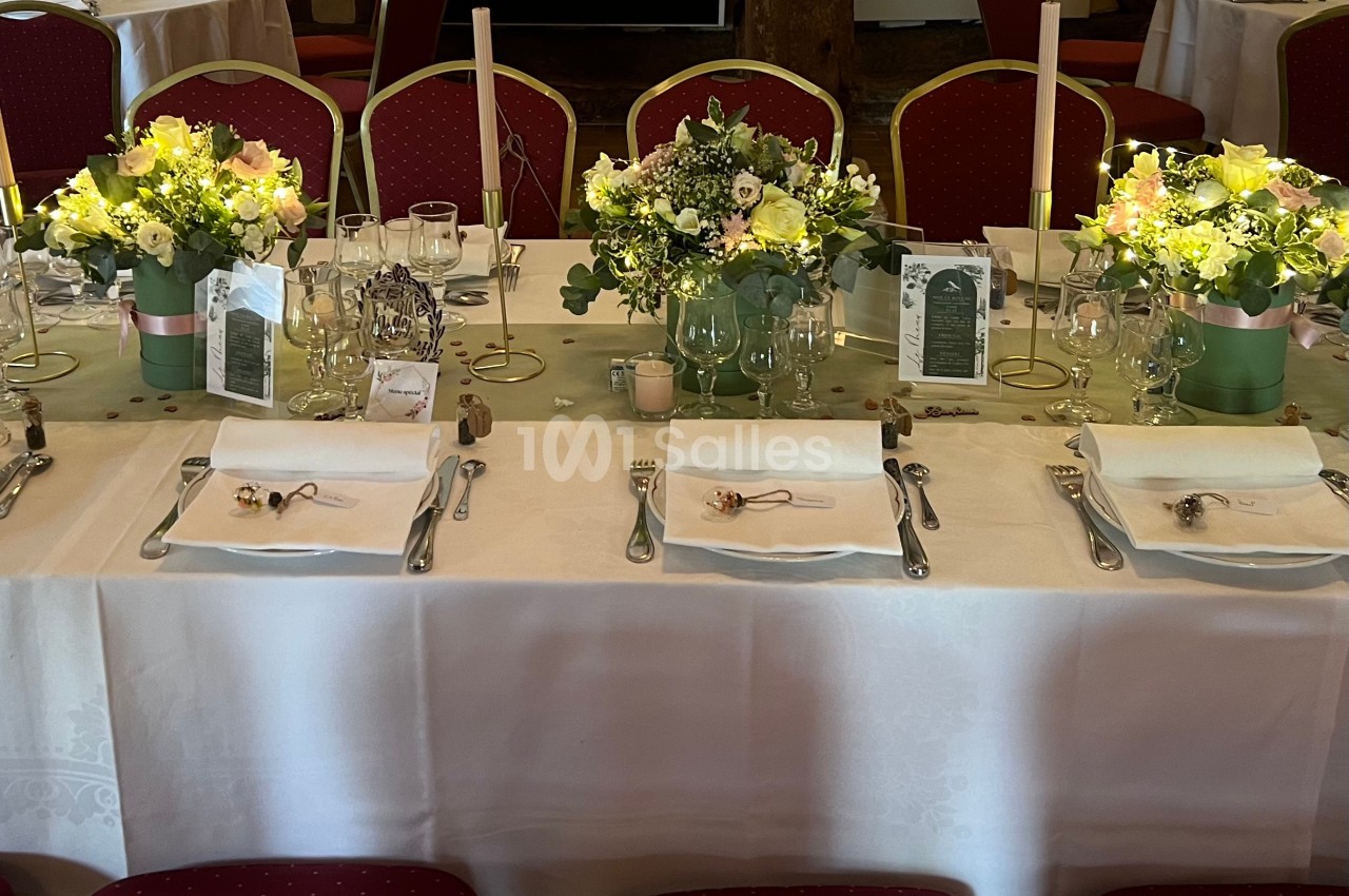 Table de réception décorée avec nappes blanches, bouquets de fleurs, bougies et couverts disposés pour un repas.