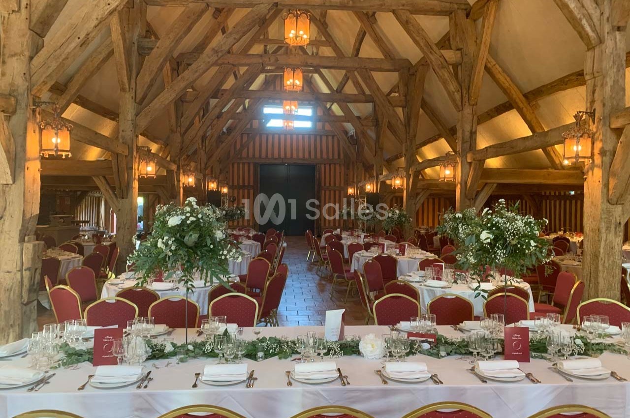 Salle de réception rustique avec poutres apparentes, tables dressées, chaises rouges et décorations florales.