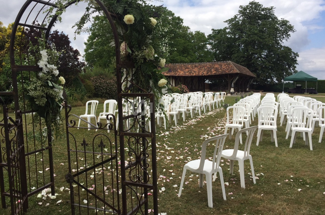 Chaises blanches alignées en extérieur pour une cérémonie, avec une arche fleurie et un chemin de pétales sur l'herbe.