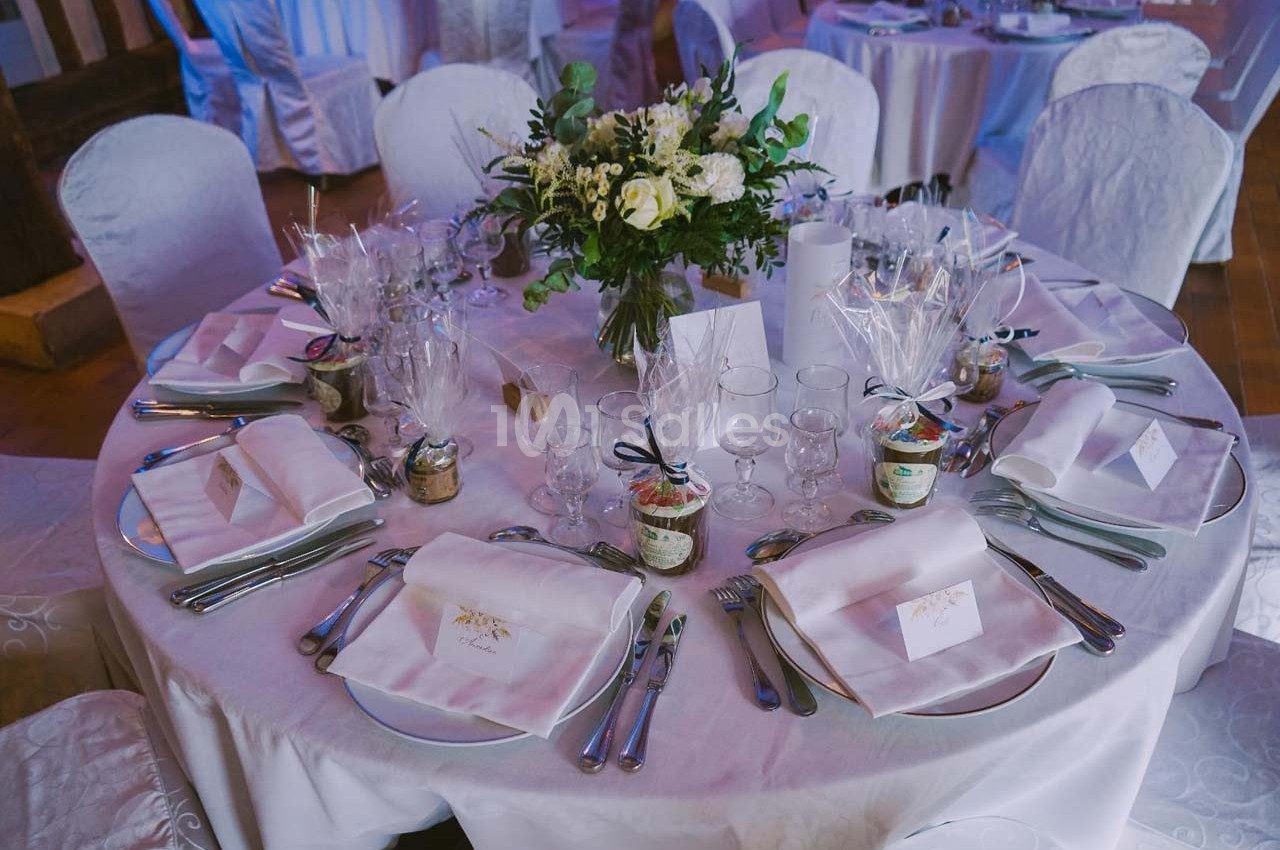 Table ronde élégamment dressée avec nappes blanches, couverts, verres, cadeaux invités et centre de table floral.