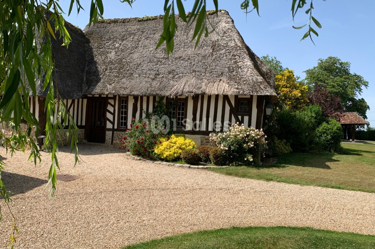 Maison à colombages avec toit de chaume entourée de fleurs et d'arbustes, située dans un jardin verdoyant.