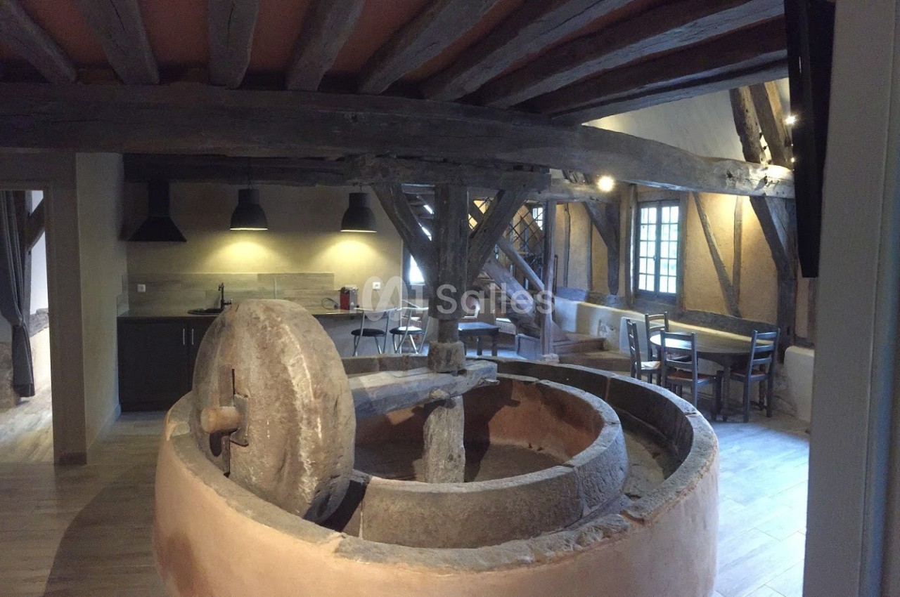 Ancien moulin en pierre intégré dans une pièce rénovée avec poutres apparentes, cuisine et coin repas.