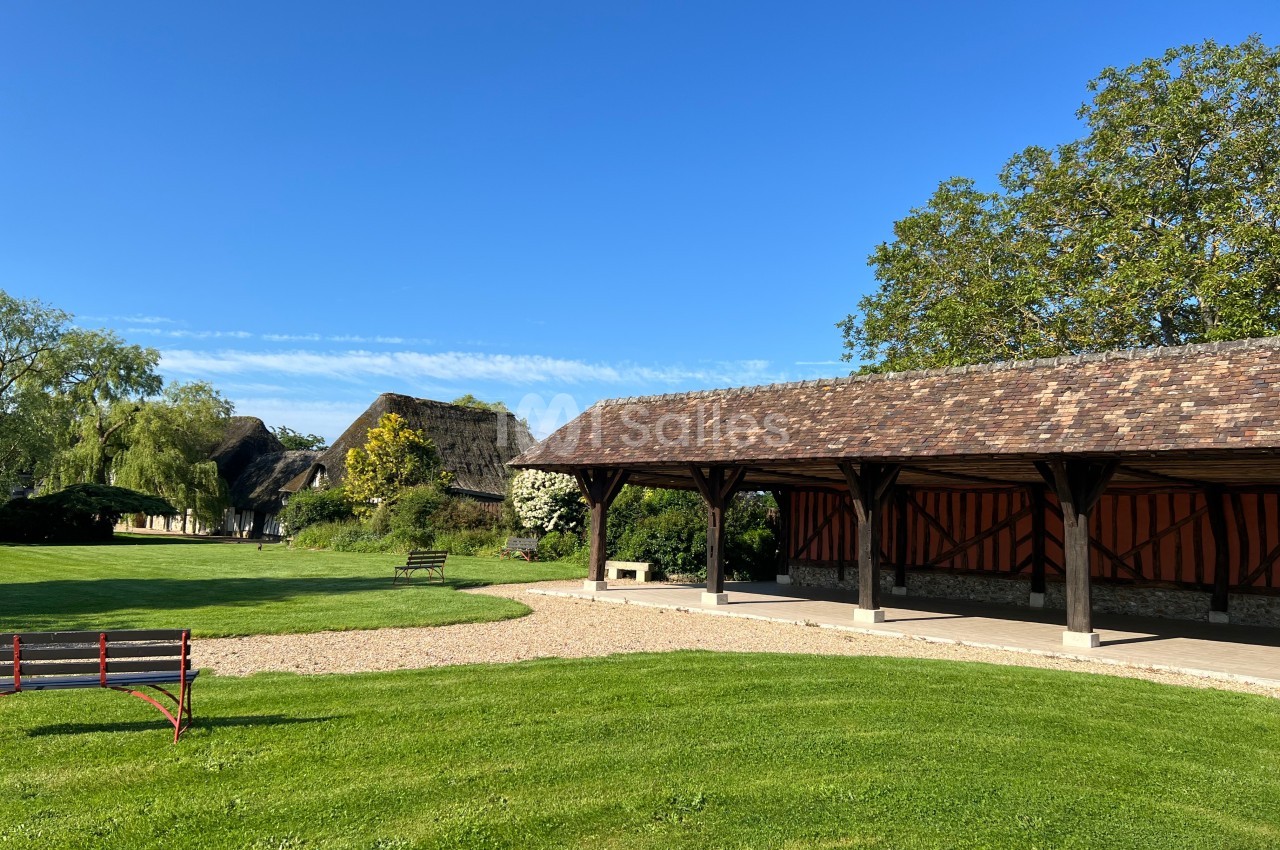 Abri en bois ouvert sur un espace vert avec des bâtiments à toit de chaume en arrière-plan sous un ciel bleu.