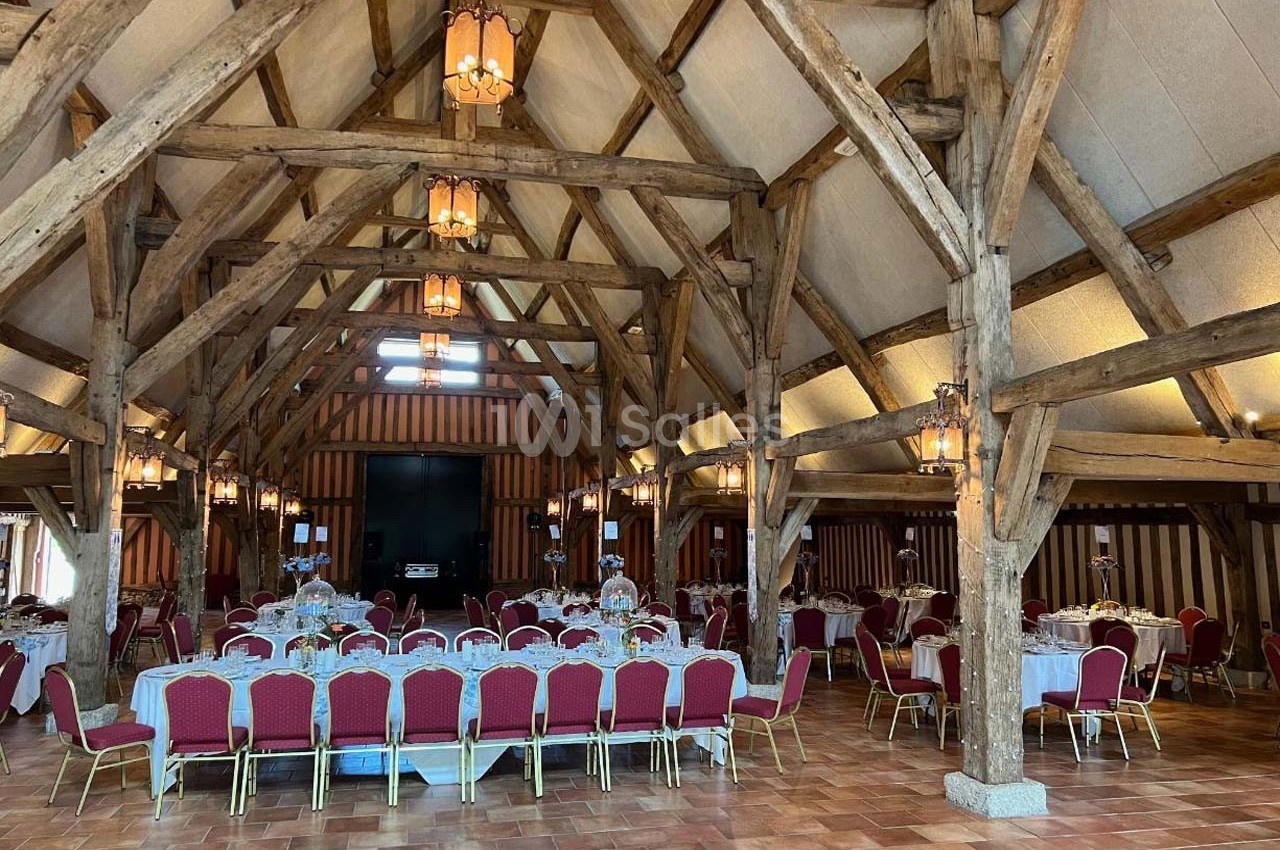 Salle de réception avec charpente en bois apparente, tables dressées et chaises rouges alignées.