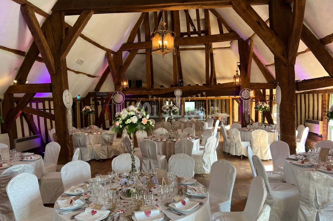 Salle de réception décorée avec des tables rondes, nappes blanches, fleurs et poutres apparentes en bois.
