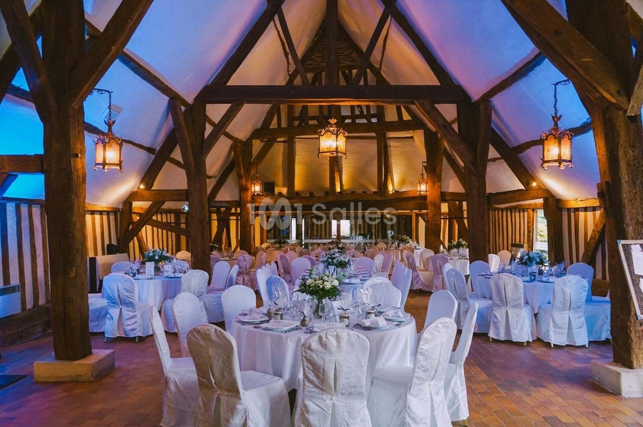 Salle de réception avec poutres apparentes, tables rondes dressées et chaises blanches, éclairée par des lanternes…