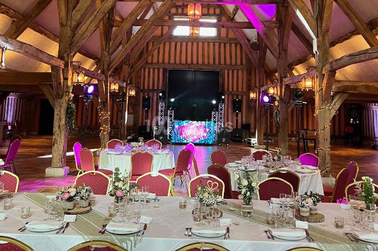 Salle de réception avec poutres apparentes, tables décorées pour un événement et éclairage coloré autour d'une scène.