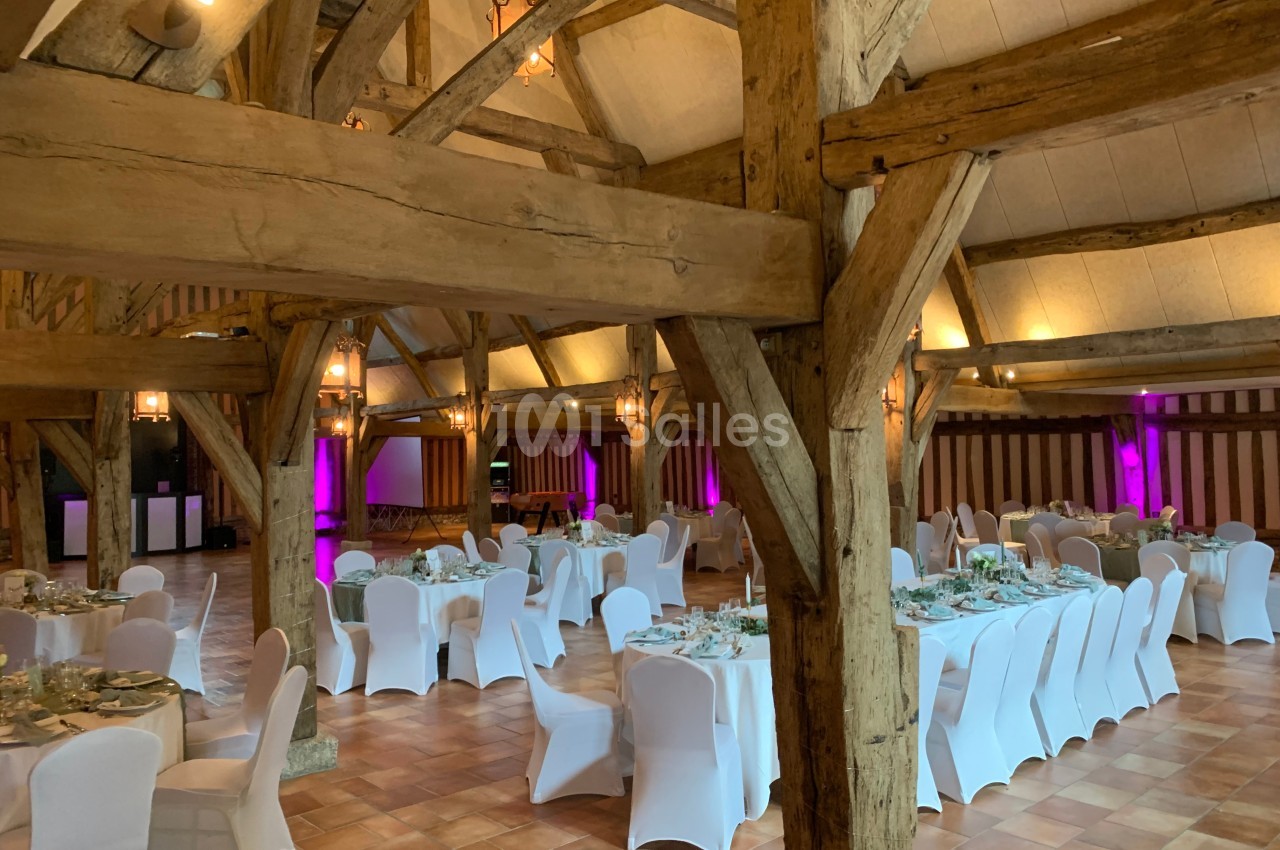 Salle de réception rustique avec poutres apparentes, tables rondes dressées et chaises blanches, éclairage tamisé.