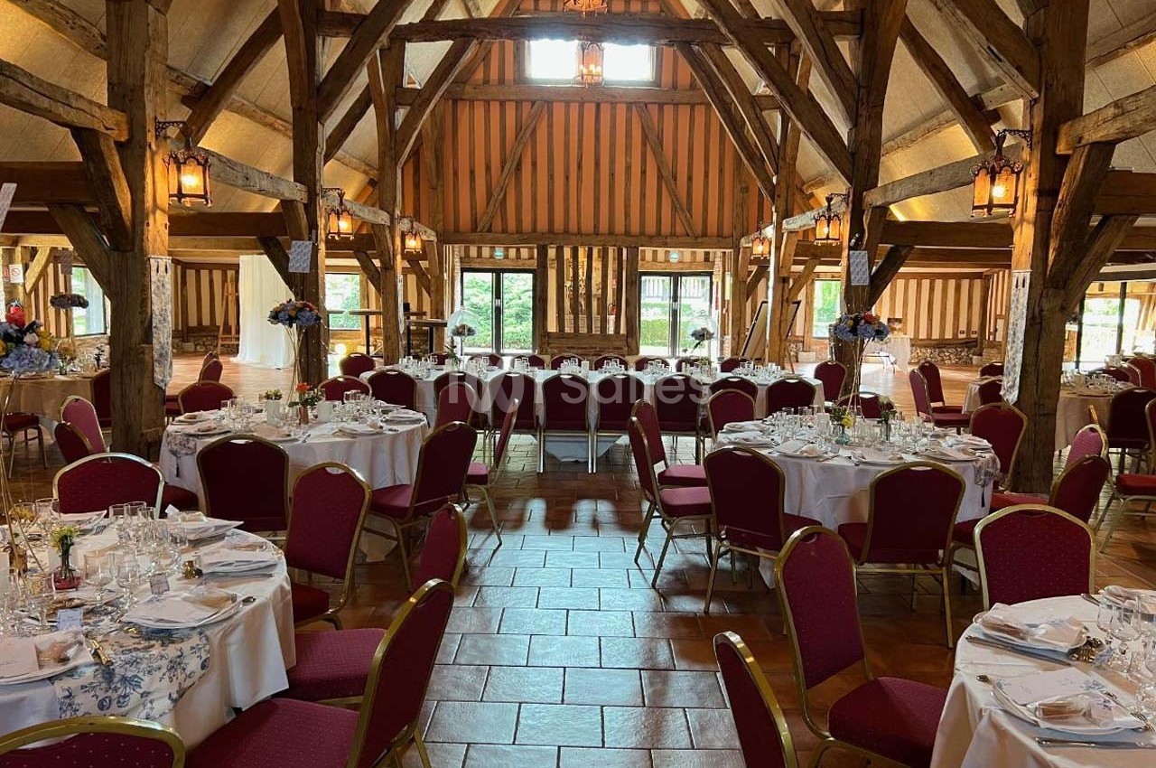 Salle de réception rustique avec poutres apparentes, tables rondes dressées et chaises rouges sur un sol en terre cuite.