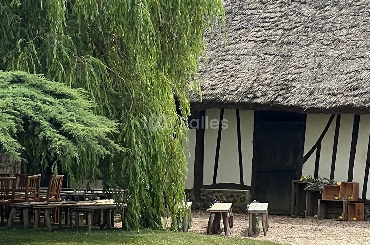 Maison à colombages avec toit de chaume, entourée d'arbres et de mobilier en bois disposé sur une pelouse.