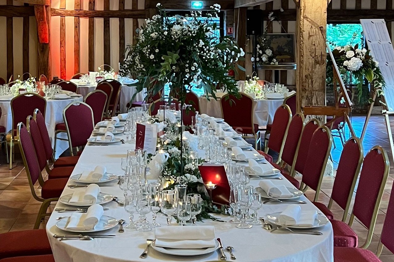 Salle de réception avec tables dressées, nappes blanches, décorations florales et chaises rouges.