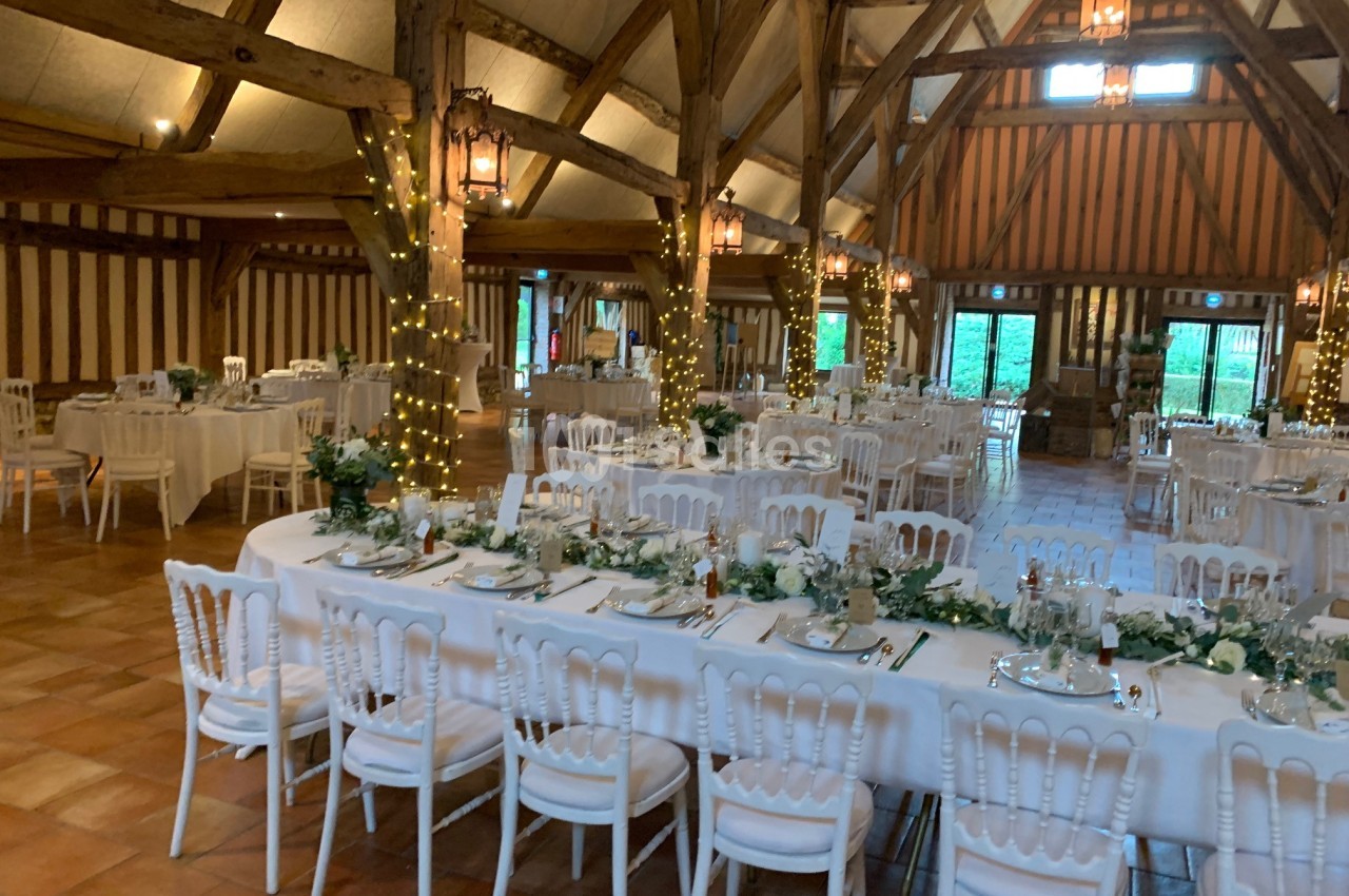 Salle de réception avec poutres apparentes, tables décorées de fleurs blanches et guirlandes lumineuses.