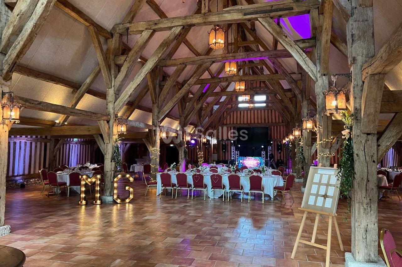Salle de réception avec charpente en bois apparente, tables dressées et décorations lumineuses.