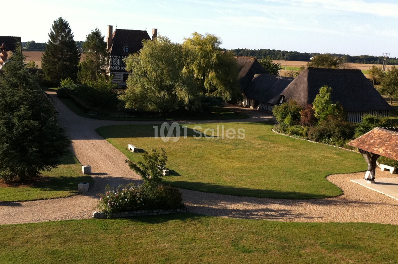 Vue d'ensemble d'un domaine avec pelouse, allées en gravier, bâtiments à colombages et toits de chaume entourés d'arbres.
