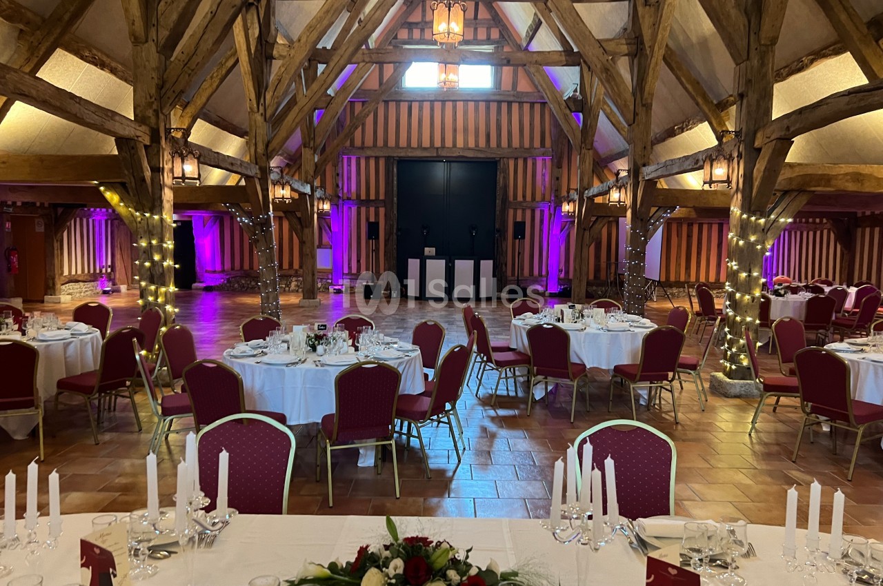 Salle de réception avec charpente en bois, tables rondes dressées et éclairages colorés sur les murs.