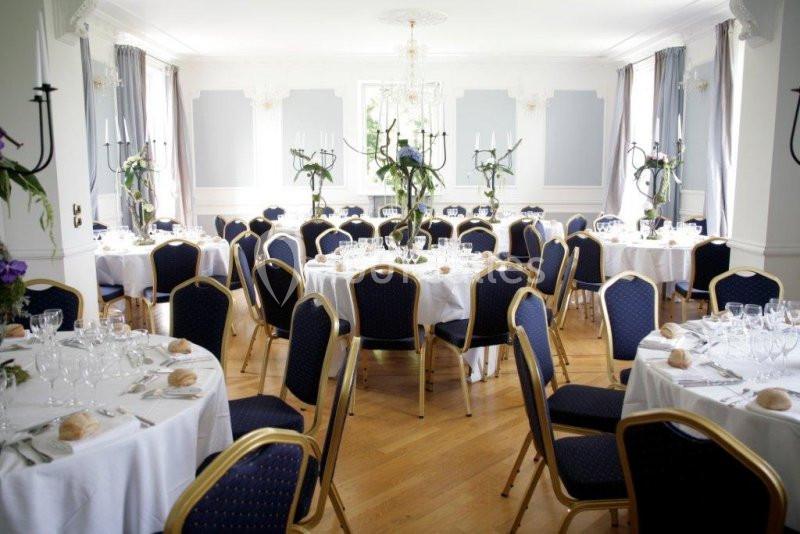 Salle de réception lumineuse avec tables rondes dressées, nappes blanches et chaises bleues entourées de décorations…