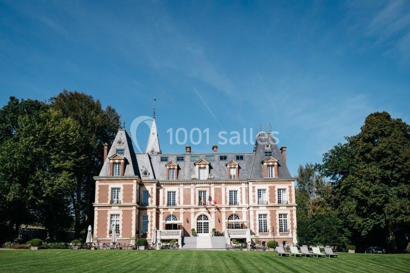 Façade d'un château en briques rouges et pierres claires, entouré de pelouse et d'arbres sous un ciel bleu.