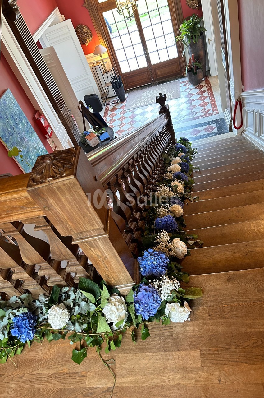 Escalier en bois décoré de fleurs blanches et bleues, menant à un hall d'entrée lumineux avec sol en carreaux.