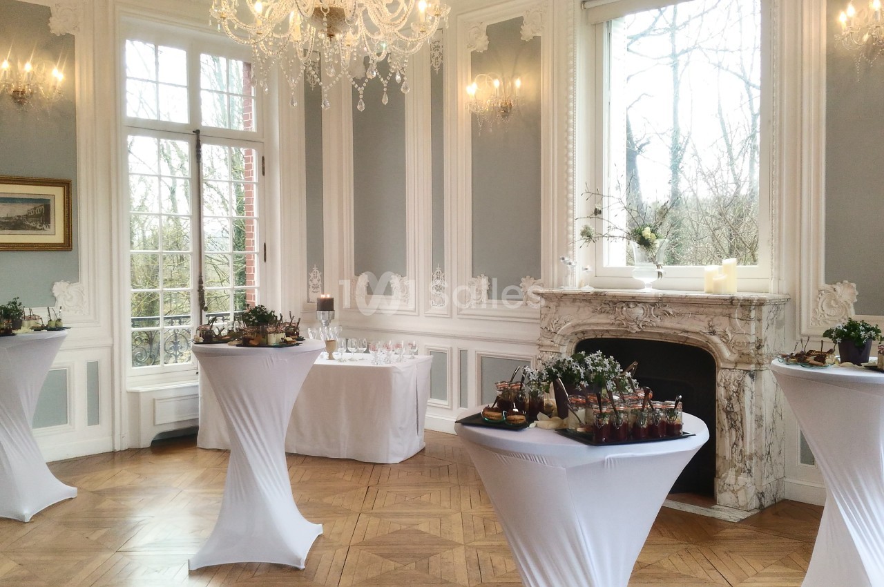 Salle élégante avec tables hautes drapées de blanc, décorée de moulures, lustre, cheminée et grandes fenêtres.