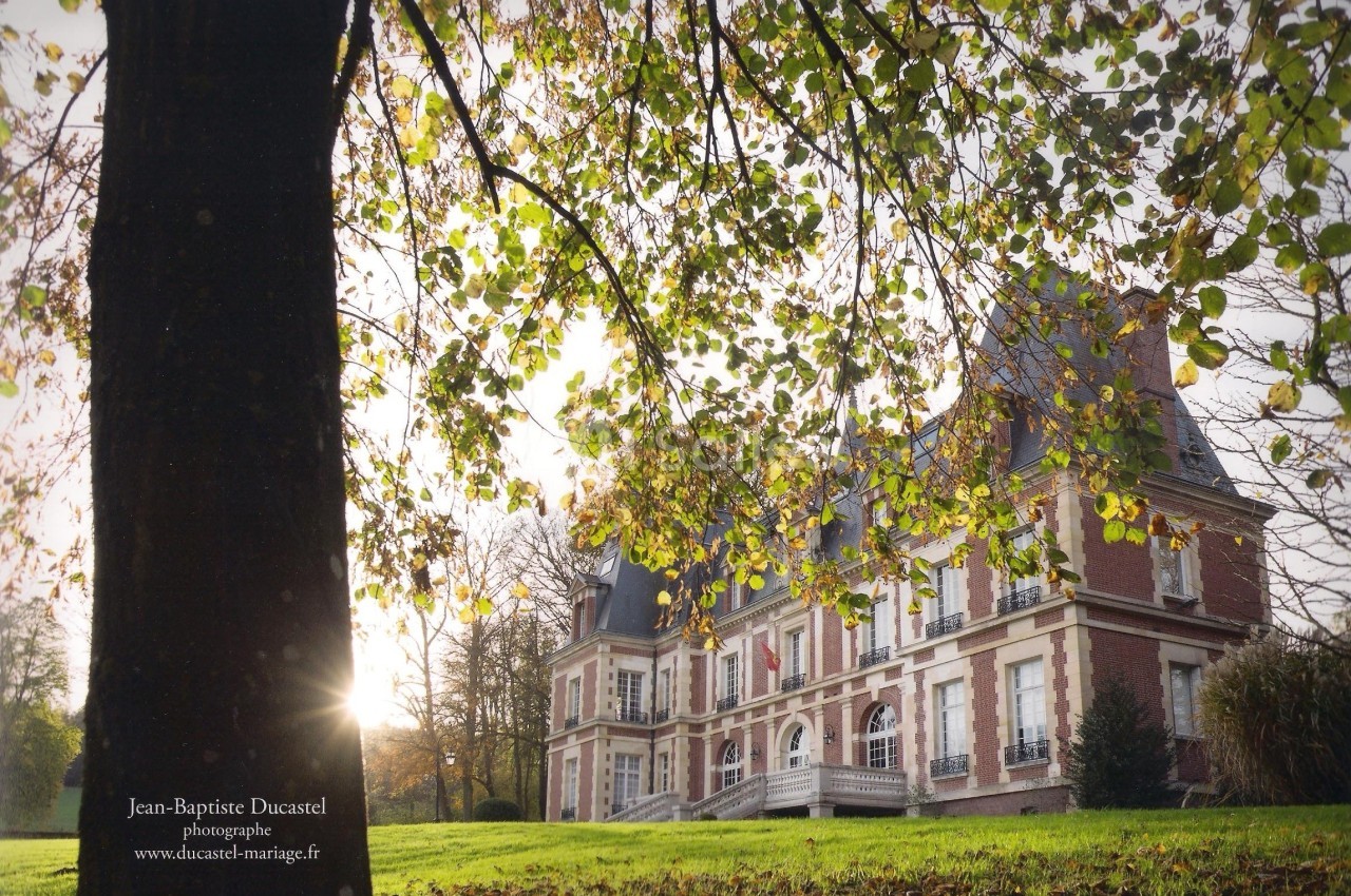 Façade d'un château en briques rouges entouré d'arbres, avec un rayon de soleil traversant les feuilles au premier plan.