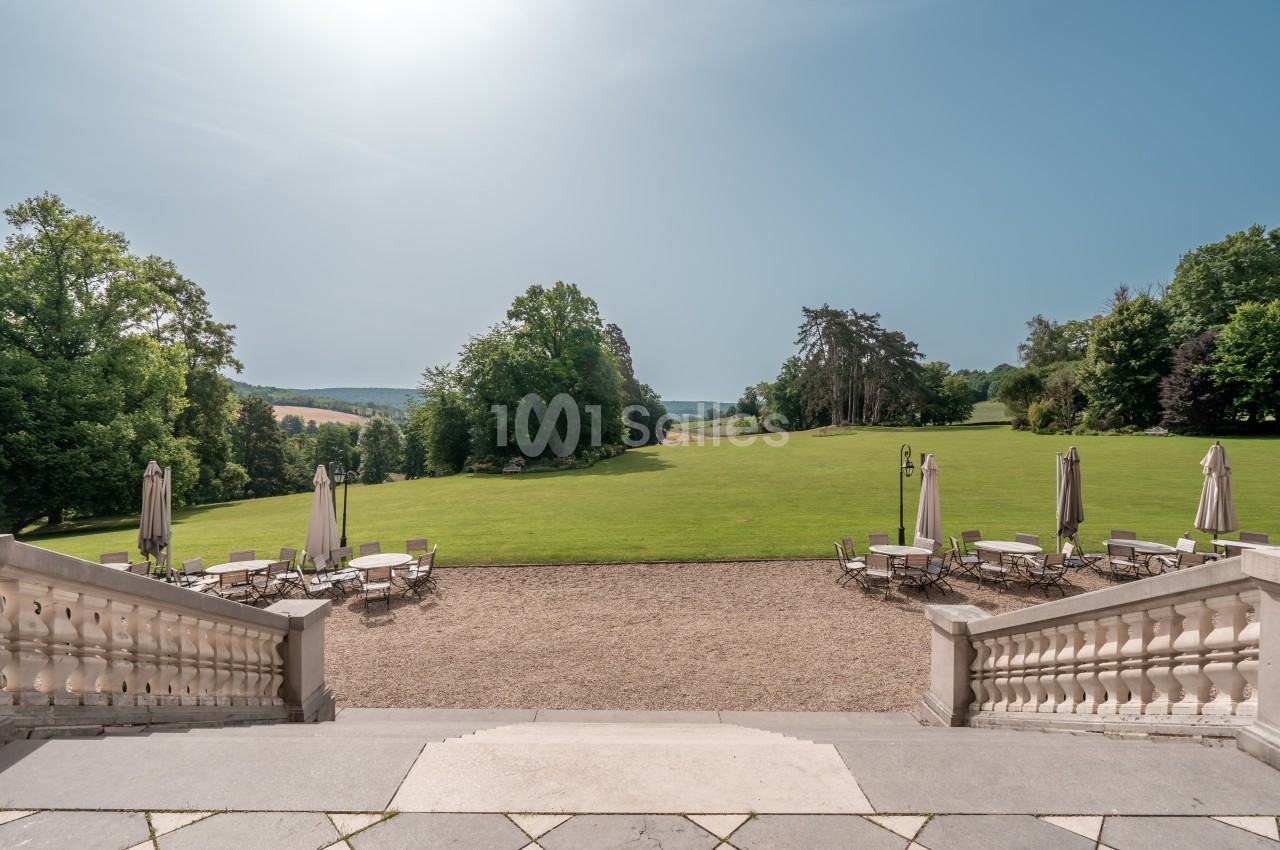 Vue depuis une terrasse sur un vaste jardin verdoyant avec des tables et parasols, entouré d'arbres sous un ciel dégagé.