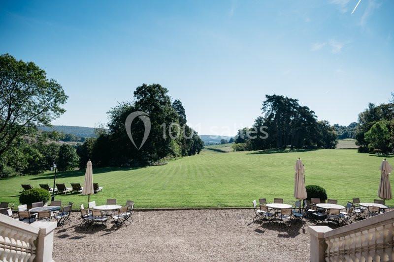 Vue sur une vaste pelouse verdoyante entourée d'arbres, avec des tables et chaises disposées en terrasse au premier plan.