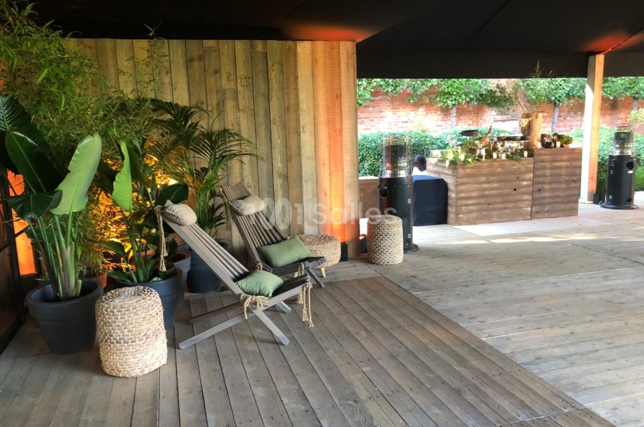 Espace extérieur avec fauteuils en bois, plantes en pots et comptoir en bois sous une structure couverte.