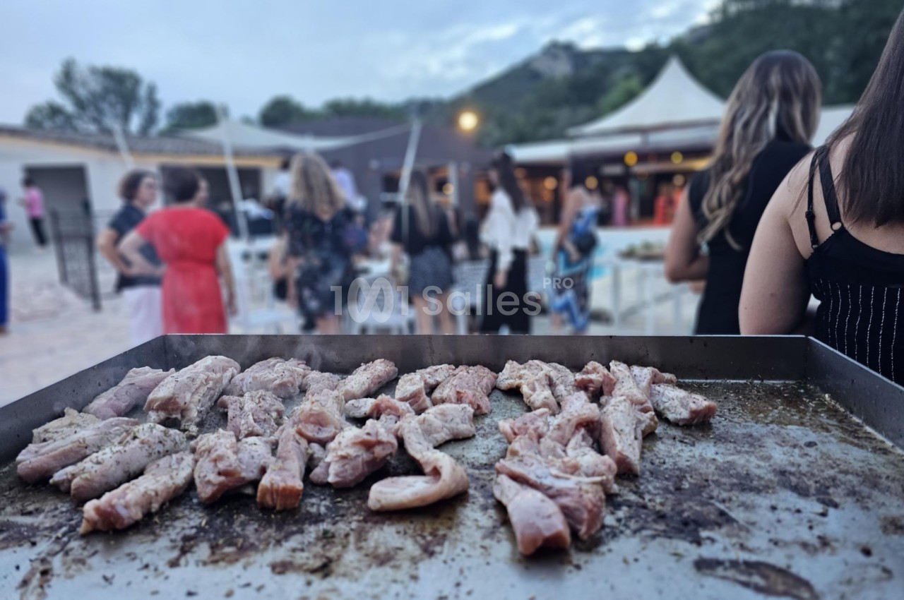 Viande en train de griller sur une plancha lors d'un événement en plein air avec des invités en arrière-plan.