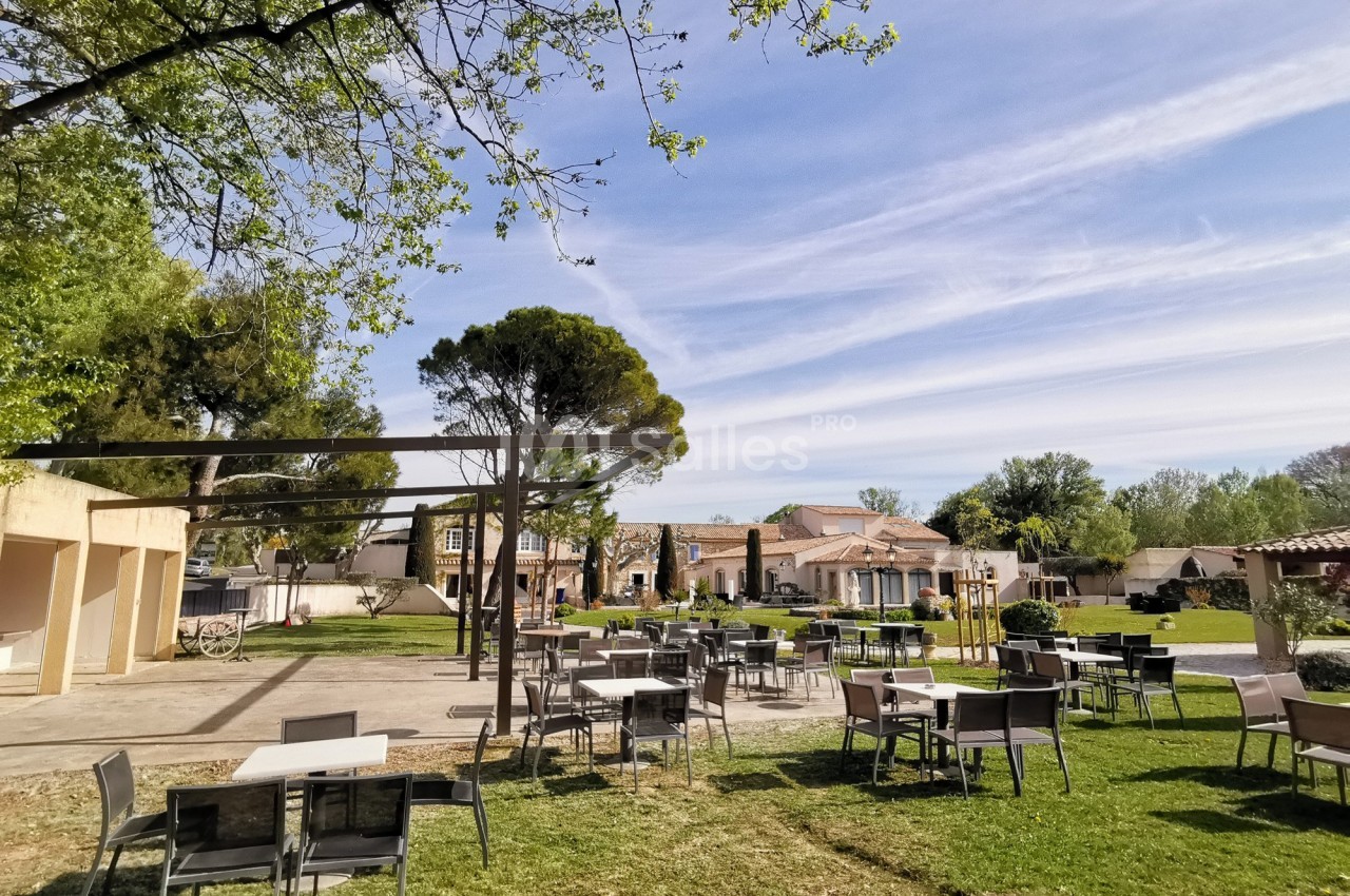 Terrasse extérieure avec tables et chaises sur une pelouse, entourée d'arbres et de bâtiments sous un ciel dégagé.