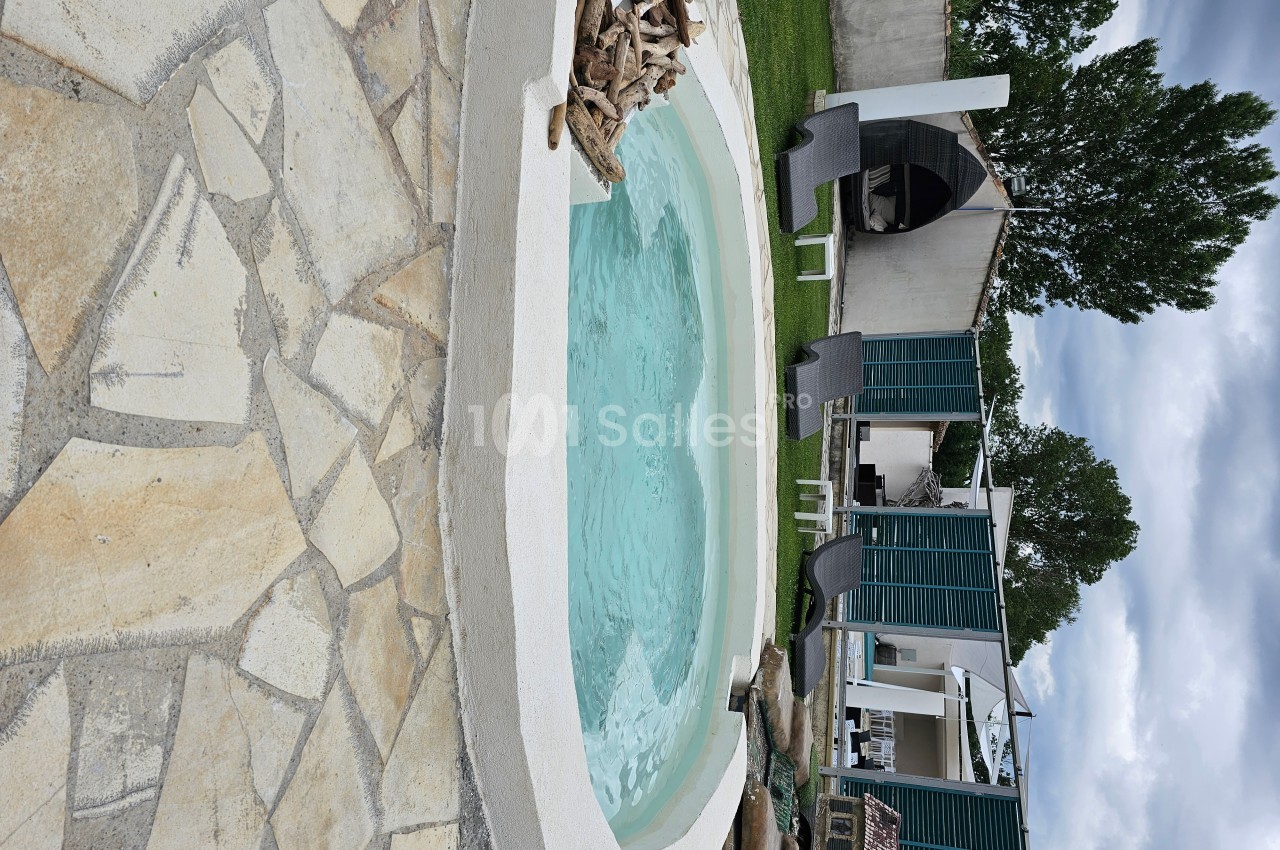Piscine extérieure ronde entourée de pierre, avec chaises longues, pelouse et abri en arrière-plan.