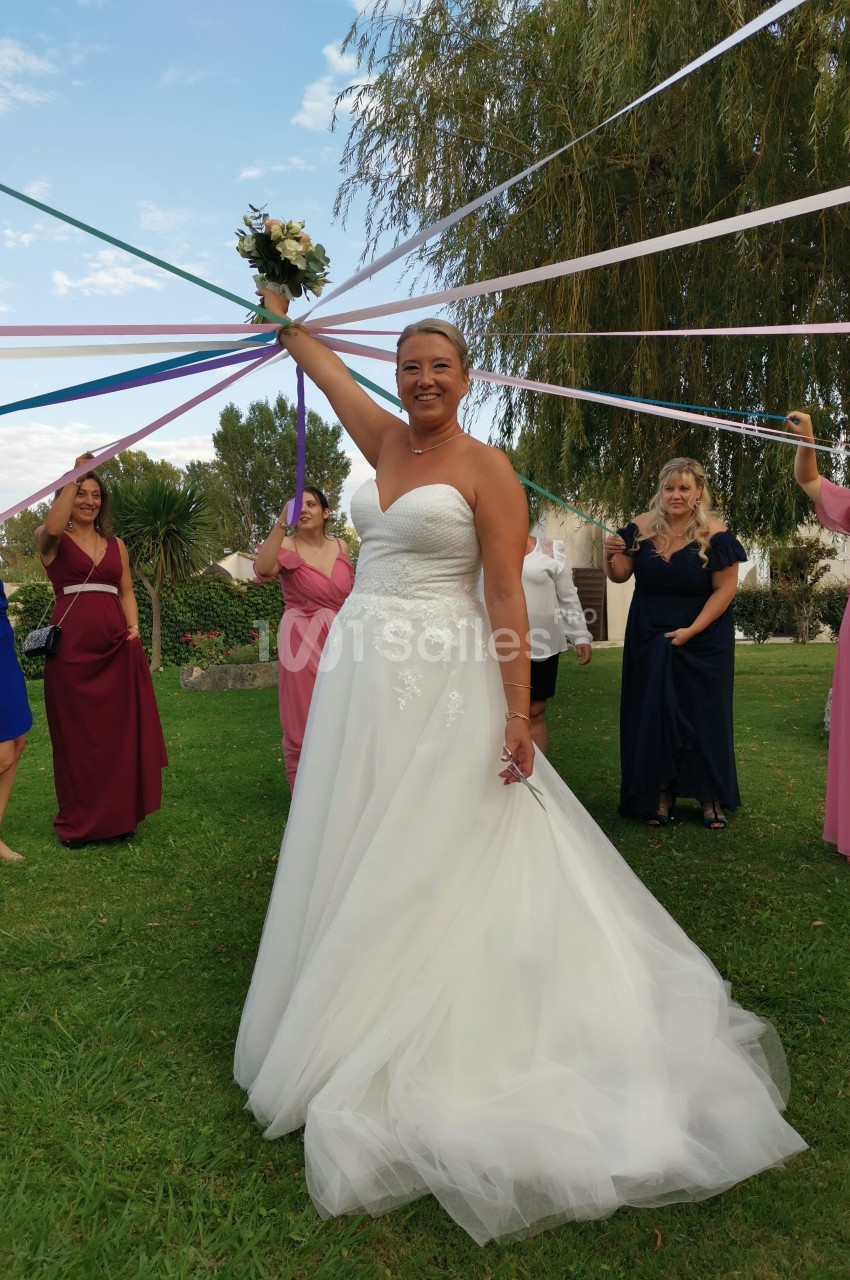 Une mariée en robe blanche tient un bouquet sous des rubans colorés, entourée d'invitées dans un jardin.
