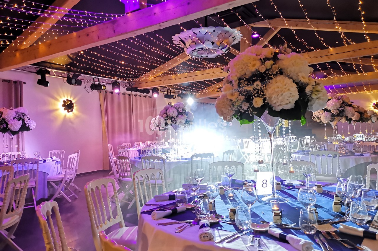 Salle de réception décorée avec des guirlandes lumineuses, des tables dressées et des centres de table fleuris.