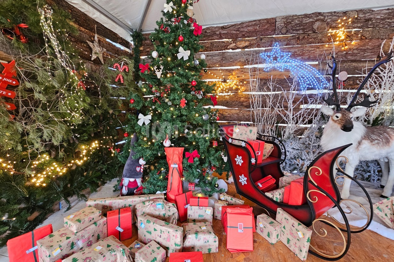 Sapin de Noël décoré entouré de cadeaux emballés, traîneau rouge et renne dans un décor festif en bois.