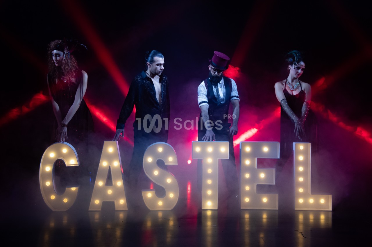 Quatre personnes posent derrière des lettres lumineuses formant ’CASTEL’, avec un éclairage rouge et une ambiance de scène.