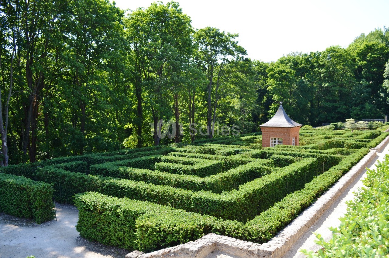 Labyrinthe de haies taillées entouré d'arbres, avec une petite cabane en briques au centre.