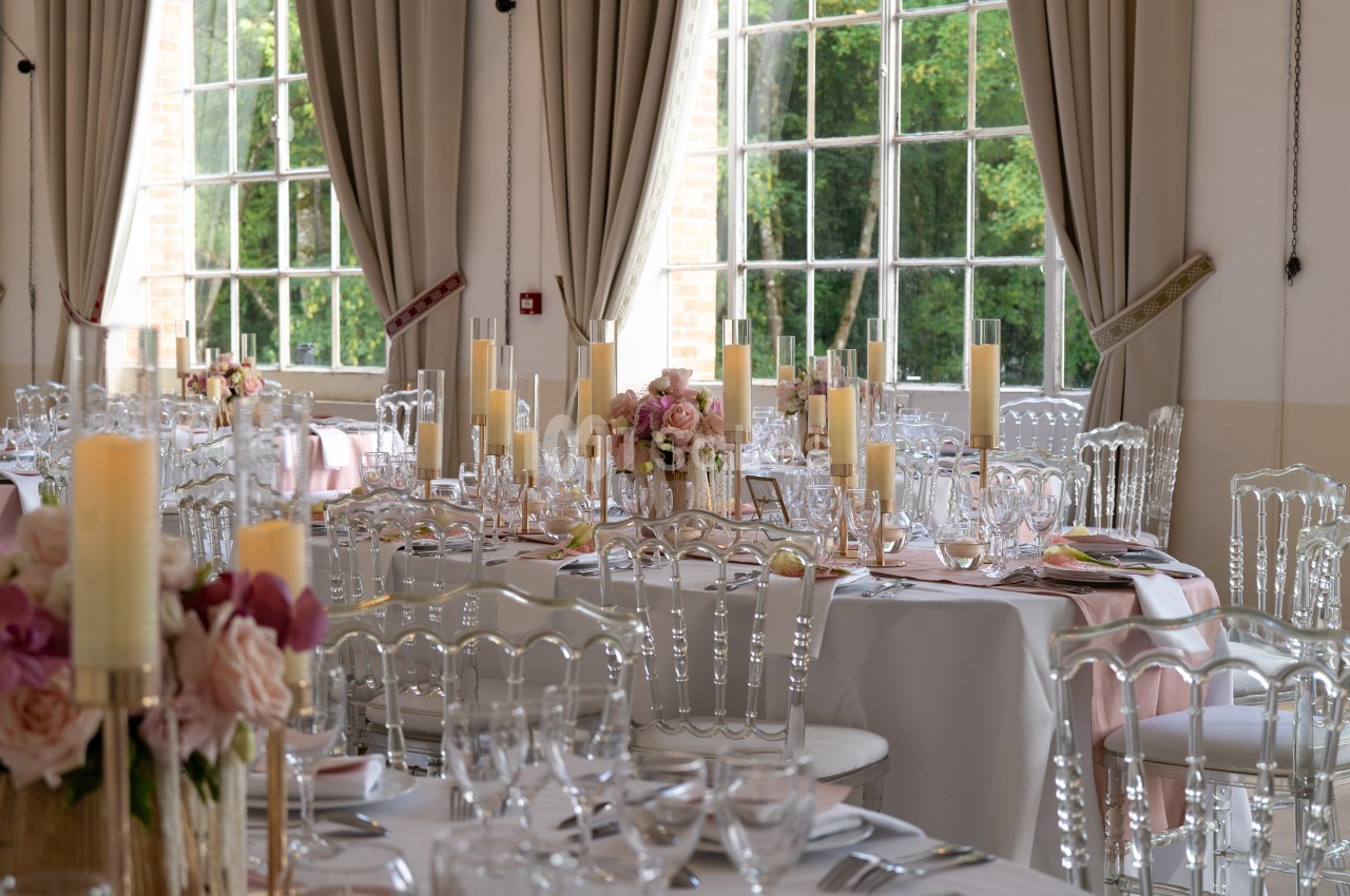 Salle de réception élégante avec tables dressées, décorations florales roses et chandeliers, près de grandes fenêtres.