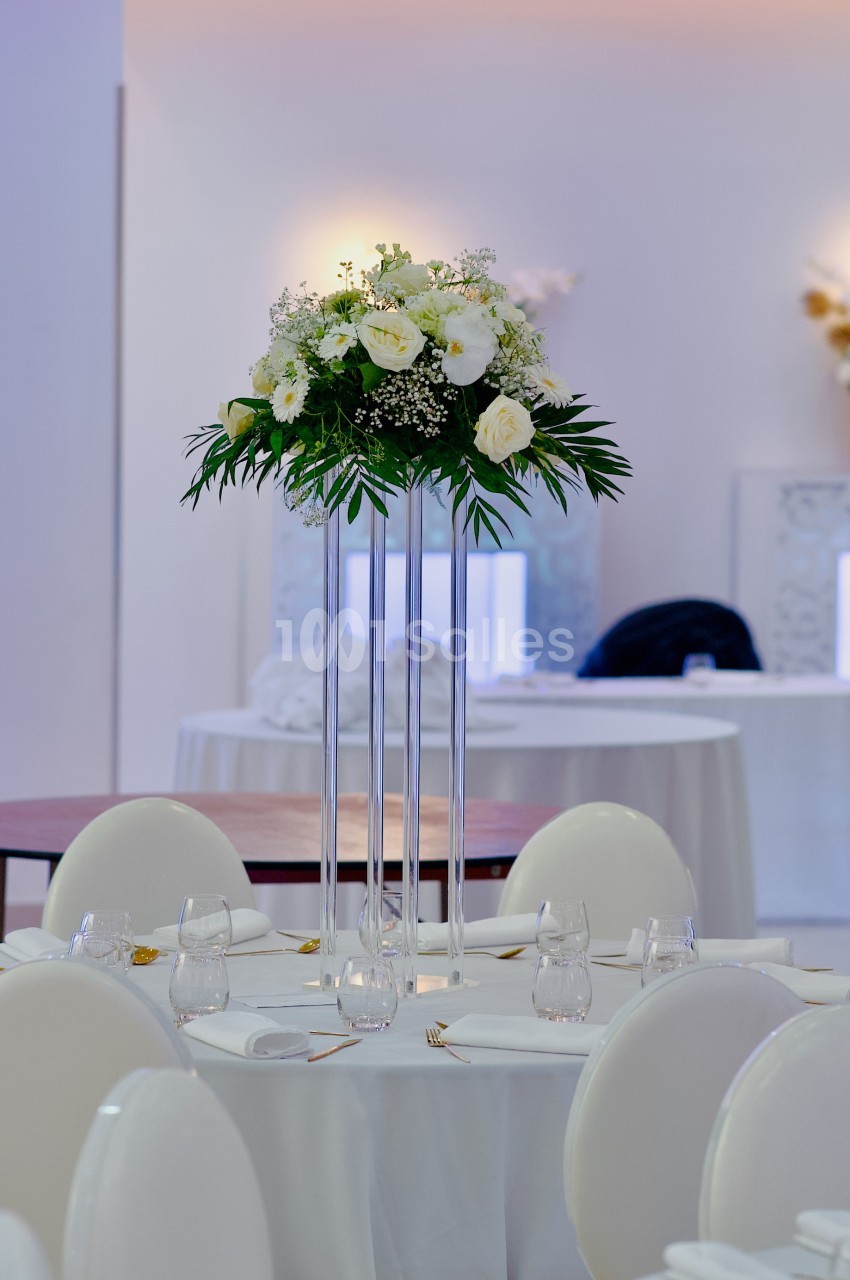 Centre de table floral avec roses blanches et feuillage, posé sur un support haut dans une salle de réception élégante.