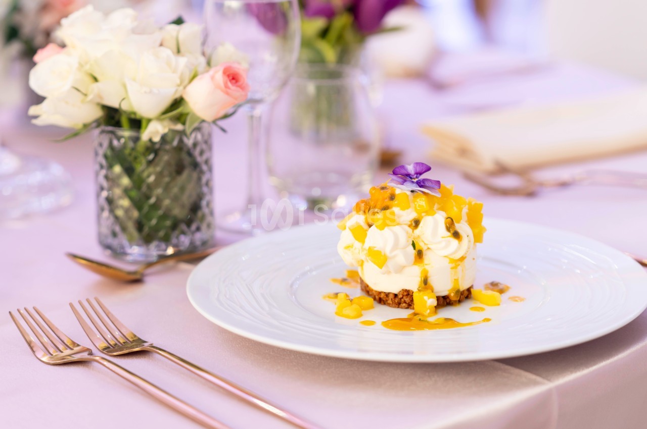 Dessert crémeux garni de morceaux de mangue et d'une fleur violette, servi sur une table élégamment dressée.