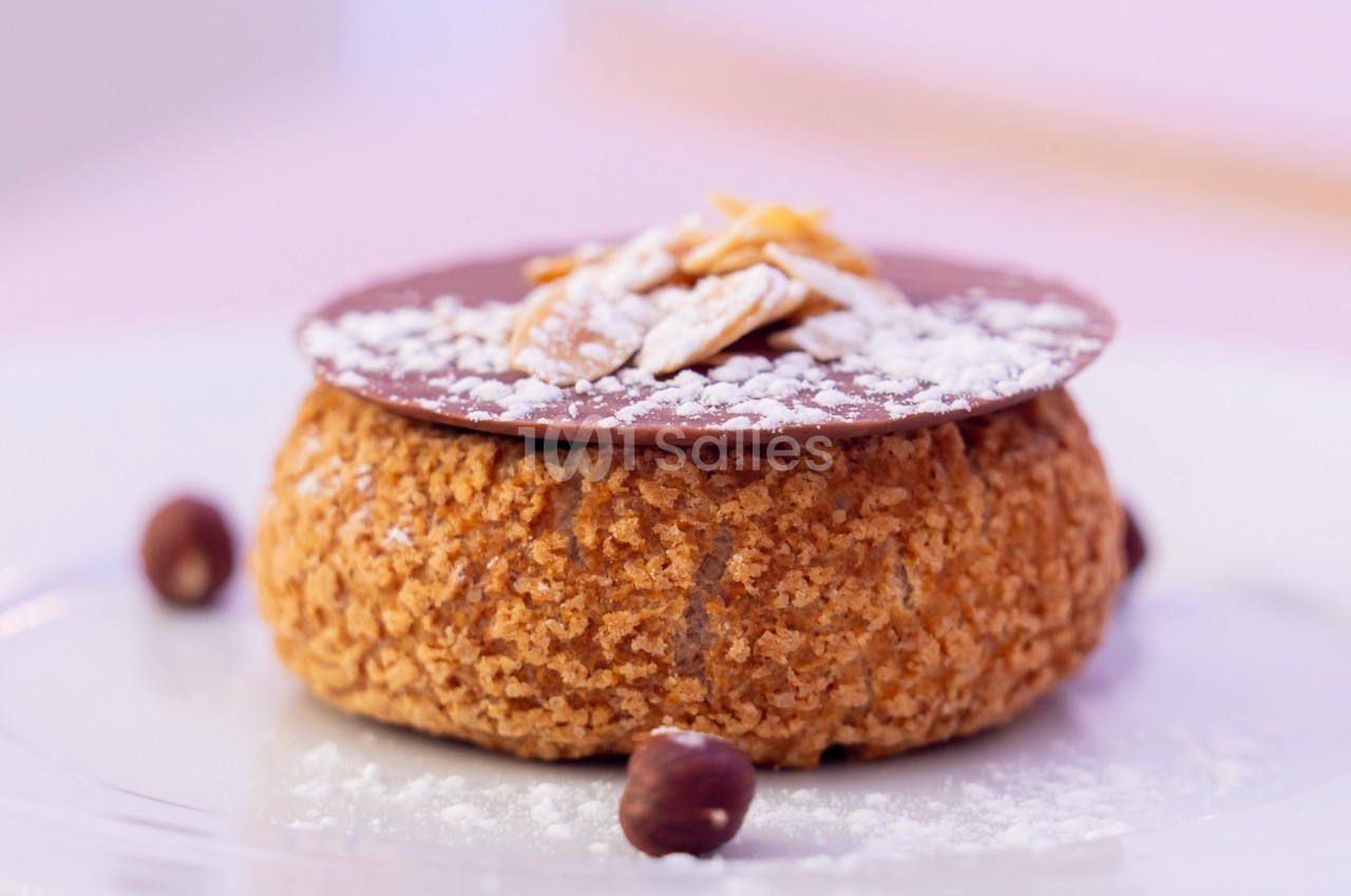 Pâtisserie garnie de crème, surmontée d'un disque de chocolat et d'éclats d'amandes, posée sur une assiette blanche.