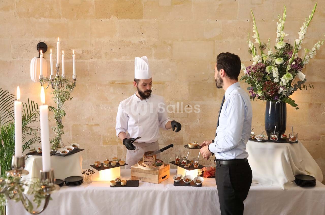 Un chef en tenue blanche prépare des plats devant un invité à une table décorée de fleurs et de chandeliers.